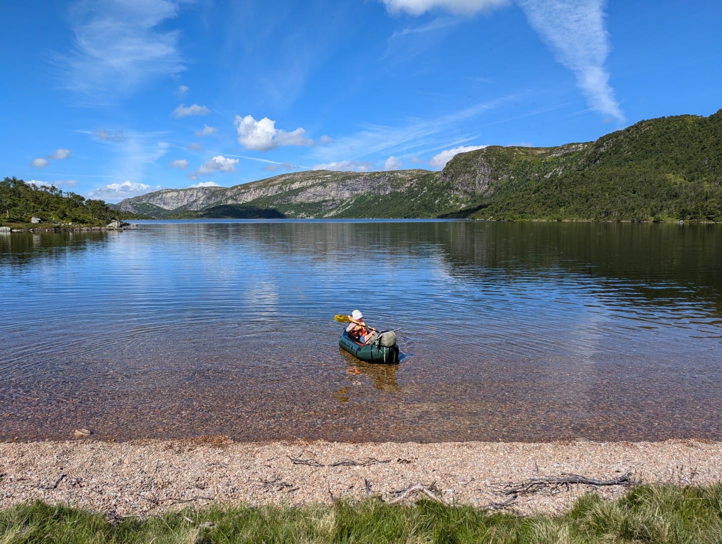 Packrafter paddling across a quiet mountain lake under a blue sky.
