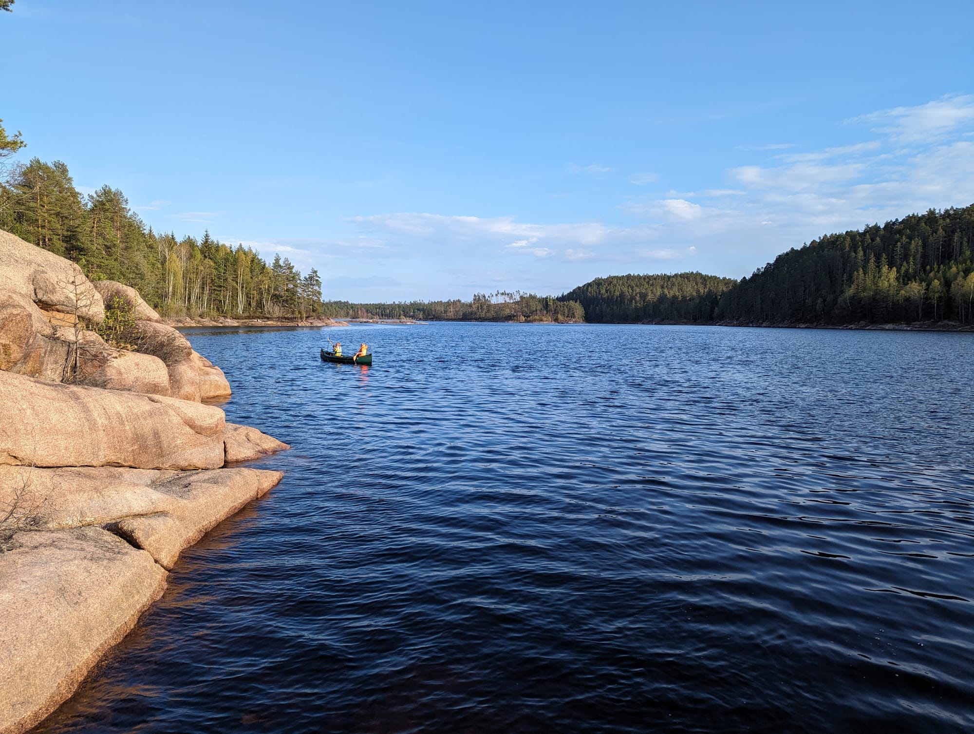Two children canoeing on a forest lake, seen from rocky shoreline under blue sky.