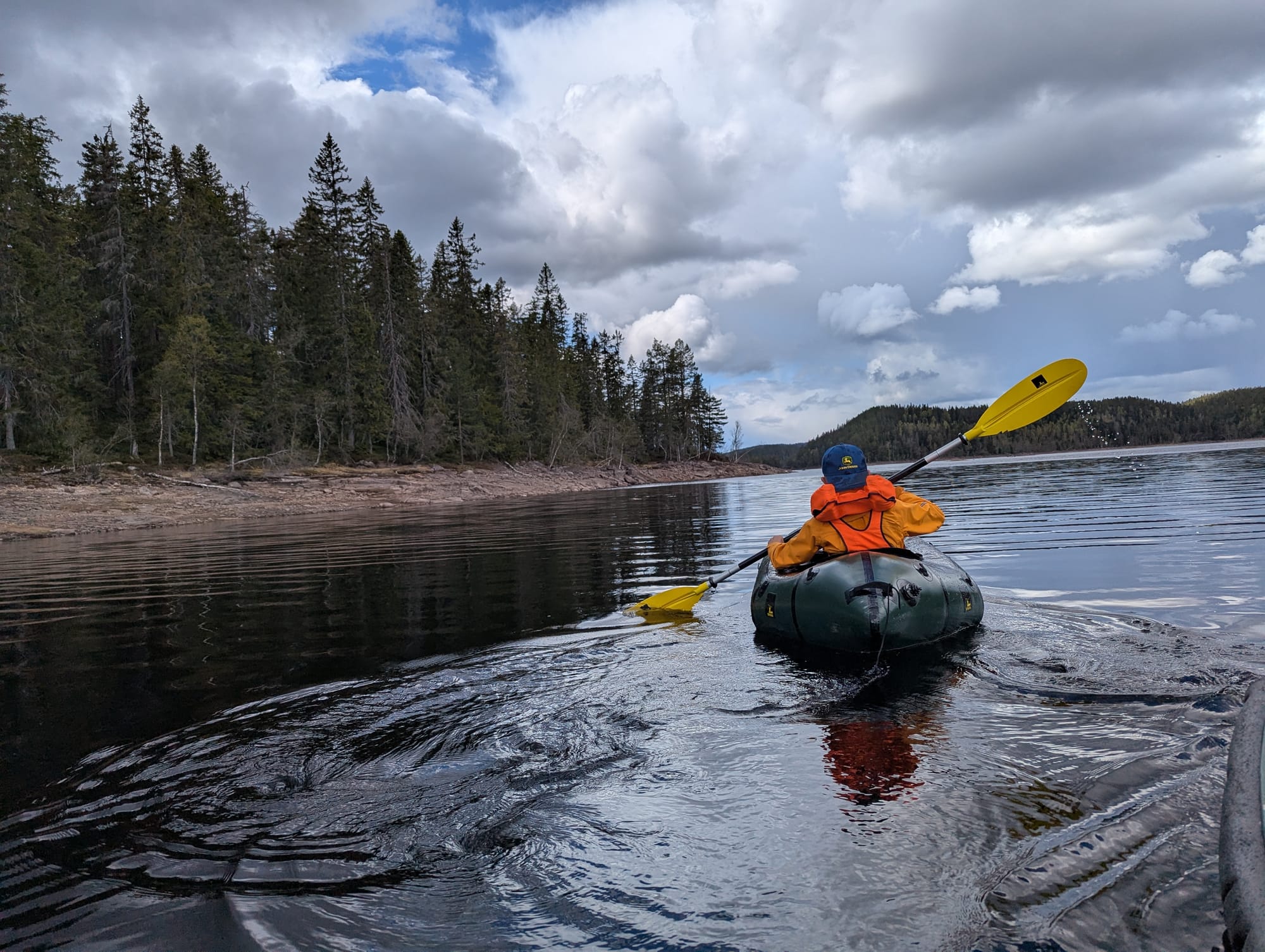 Child in an orange jacket paddling a green packraft along the shoreline of a forested lake, heading toward camp under a cloudy spring sky.
