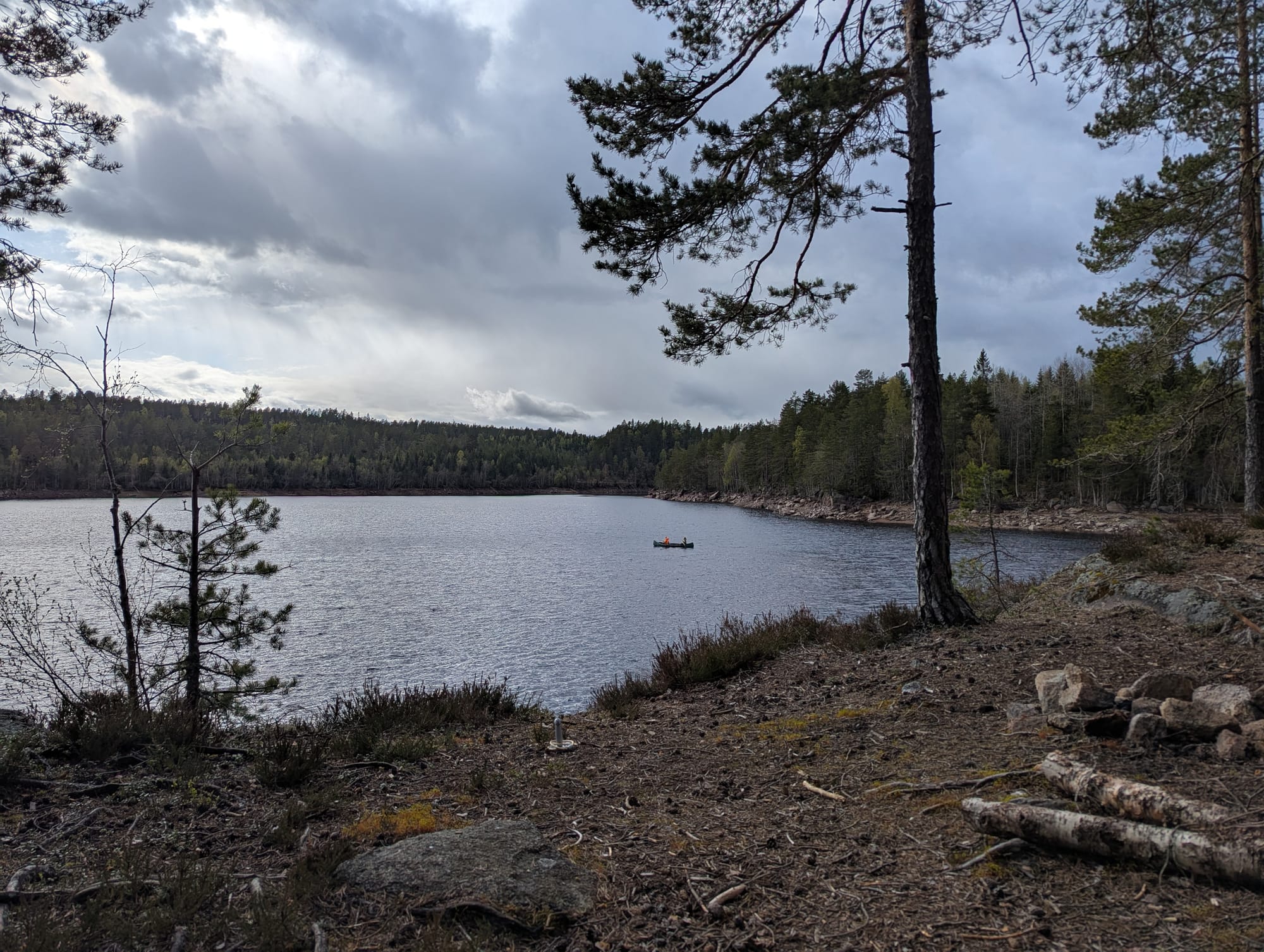 View from a forested lakeshore campsite, looking across a calm lake where two children in a canoe are trolling for fish under a cloudy spring sky.